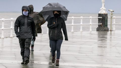 Un grupo de personas pasea por la playa de San Lorenzo de Gijon, bajo la lluvia y el viento