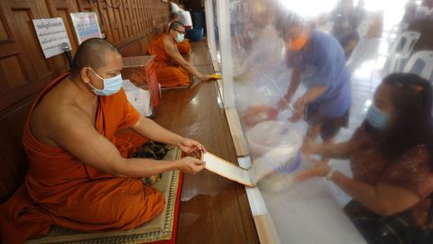Monjes budistas, protegidos con mascarillas y una cortina de plástico reciben ofrendas de sus devotos en un monasterio de Bangkok, Tailandia