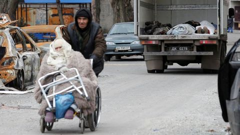 Un hombre empuja una silla de ruedas con una anciana y, al fondo, un camión con cadáveres en Mariúpol