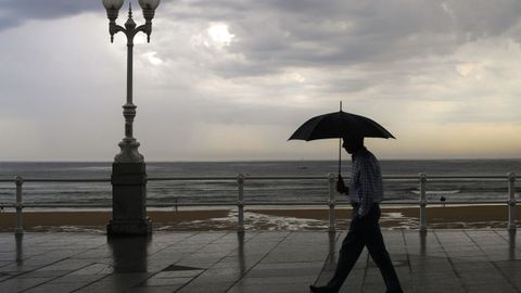 Un hombre se protege con el paraguas de la lluvia, en el paseo de muro de la playa de San Lorenzo de Gijón, en una imagen de este verano