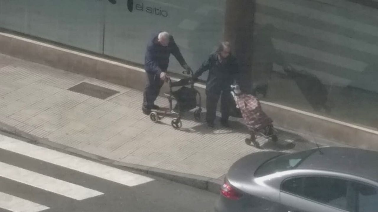 Una pareja de ancianos, con un carrito de la compra y un andador, por una calle de Gijón, durante esta crisis sanitaria