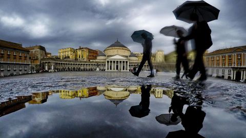 Varias personas se reflejan en un charco mientras se protegen de la lluvia con paraguas en el centro de Nápoles
