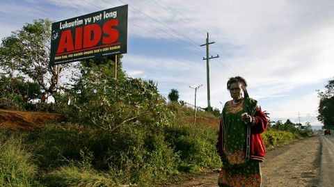 Una mujer camina descalza sobre el asfalto en Papa Nueva Guinea; en el fondo, un cartel que alerta de la amenaza del virus del SIDA.
