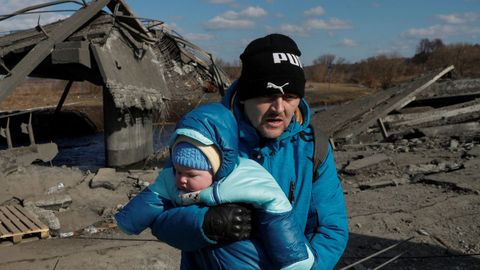 Un hombre lleva a un bebé mientras cruza el río junto a un puente destruido durante la evacuación de Irpin