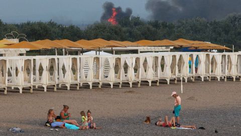 La gente descansa en una playa mientras el humo y las llamas se elevan después de las explosiones en una base aérea militar rusa, en Novofedorivka, en Crimea.  