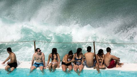 Varias personas permanecen sentadas en el borde de la piscina Icebergs junto al mar mientras una ola se eleva gracias al viento en la playa de Bondi, en Sidney.